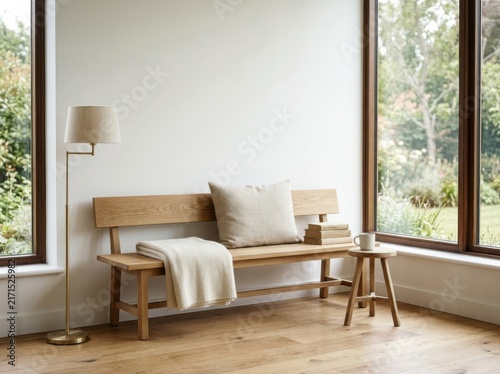 Minimalist interior featuring a solid oak bench with a neutral linen cushion and knit throw, accompanied by a brass floor lamp and a wooden stool with a mug and books by a large window.