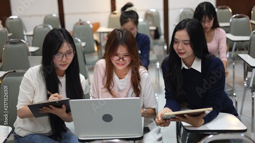 Asian university students collaborating on group project with digital tablet and laptop in classroom hall. Modern higher education, academic learning, Gen Z lifestyle and study teamwork concept.