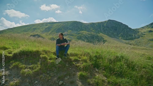 Man sits on grassy hill while looking at mountain landscape under bright sky with clouds