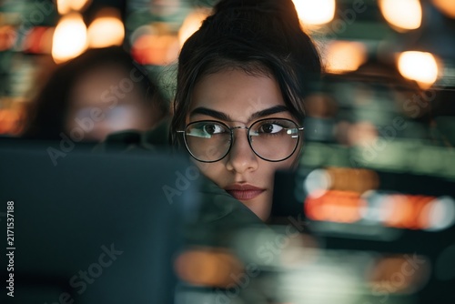 Close up of a young woman wearing glasses looking at a computer screen in a dark office space
