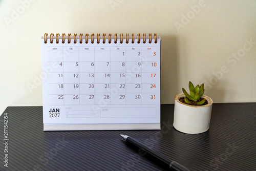 Spiral desk calendar showing January 2027 standing on a dark textured desk next to a potted succulent and a pen. Minimalist office planning setup.