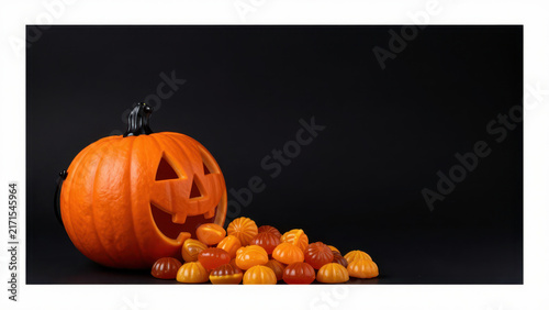 Halloween pumpkin jack o lantern carved face beside colorful candy pile on black background, creating playful seasonal scene for holiday greeting and celebration design