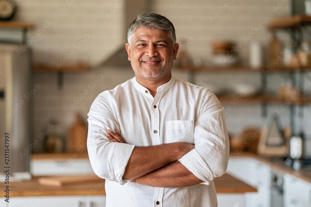 Obraz premium Middle-aged indian man standing with arms crossed smiling in a modern home kitchen