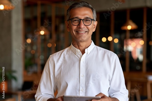 Smiling mature businessman wearing glasses and white shirt holding digital tablet inside office