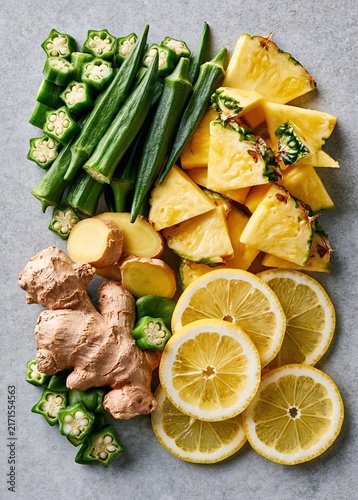 Fresh okra, pineapple chunks, ginger root and lemon slices on a grey stone background