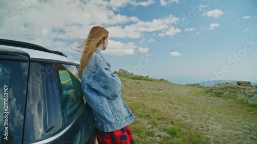 Woman stands by car and looks at mountains on a sunny day in nature with clouds in the sky