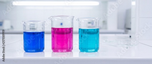 Three glass laboratory beakers filled with vibrant blue magenta and cyan chemical liquid solutions on sterile white science workbench