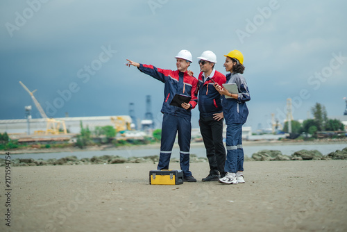 Three marine engineers pointing toward industrial port while discussing vessel inspection, maintenance planning and shipyard operations near shoreline.