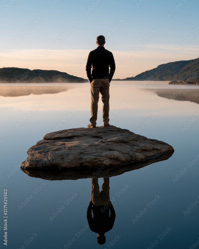 Obraz premium Lone person stands on rock by calm lake, surrounded by mountains, reflecting in water, peaceful sunrise, solitude, nature, outdoor