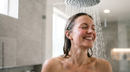 an adult woman happily taking a shower, leaning forward toward a rainfall shower stream, wet hair and realistic water droplets, personal care and wellness advertising concept