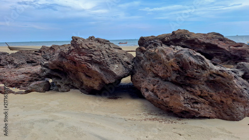 Foto Close perspective of two heavy porous rock boulders sitting on soft beach sand with a distant wooden fishing boat on the calm ocean horizon