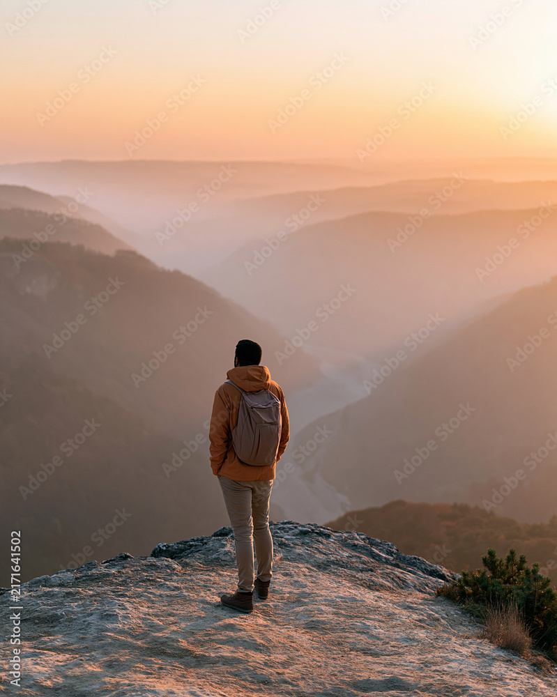 Fototapeta premium One person stands atop a rocky outcrop at dawn. Golden light bathes distant valleys and rolling hills. Steam rises gently from the valley floor. Backpack rests on their shoulders