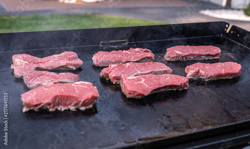 Fresh beef steaks cooking on a hot outdoor flat top griddle, with smoke rising during barbecue preparation.