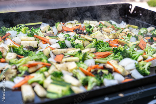 Mixed fresh vegetables cooking on a hot outdoor flat-top griddle, including broccoli, zucchini, carrots, onions and mushrooms, with steam rising.