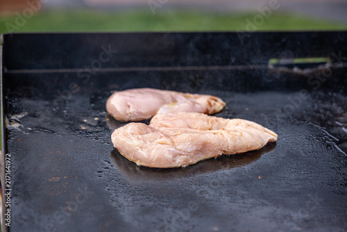 Chicken breast fillets cooking on a hot outdoor hot top griddle, with steam rising from the meat during barbecue preparation.