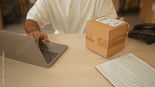 Man holds parcel with one bare hand while typing on laptop at a building desk surrounded by boxes and order notebook; entrepreneurial focus.