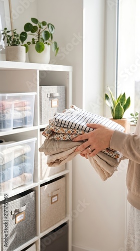 Woman holding stack of clean folded clothes near storage boxes on shelf at home. Laundry chores and wardrobe organization concept. Seasonal summer clothes storage and tidying.