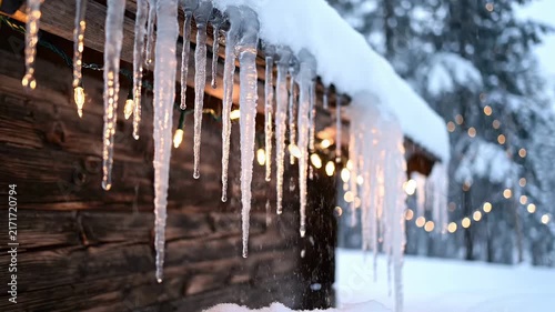 Icicles hanging from a snow covered roof with festive lights in the background.