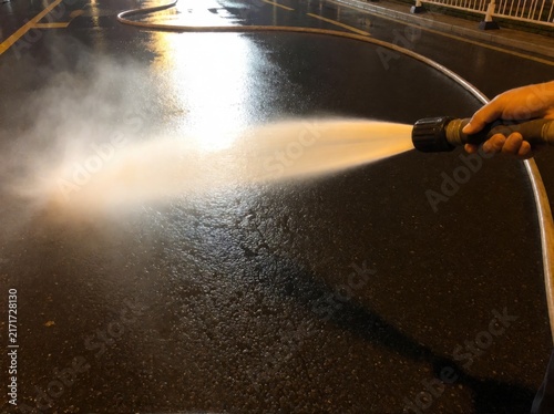 A close-up view of a person using a pressurized hose to spray water onto a dark asphalt street surface.