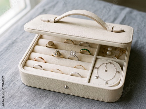 Open beige leather jewelry box displaying gold rings, a diamond solitaire, bracelet, and pearls on gray fabric.