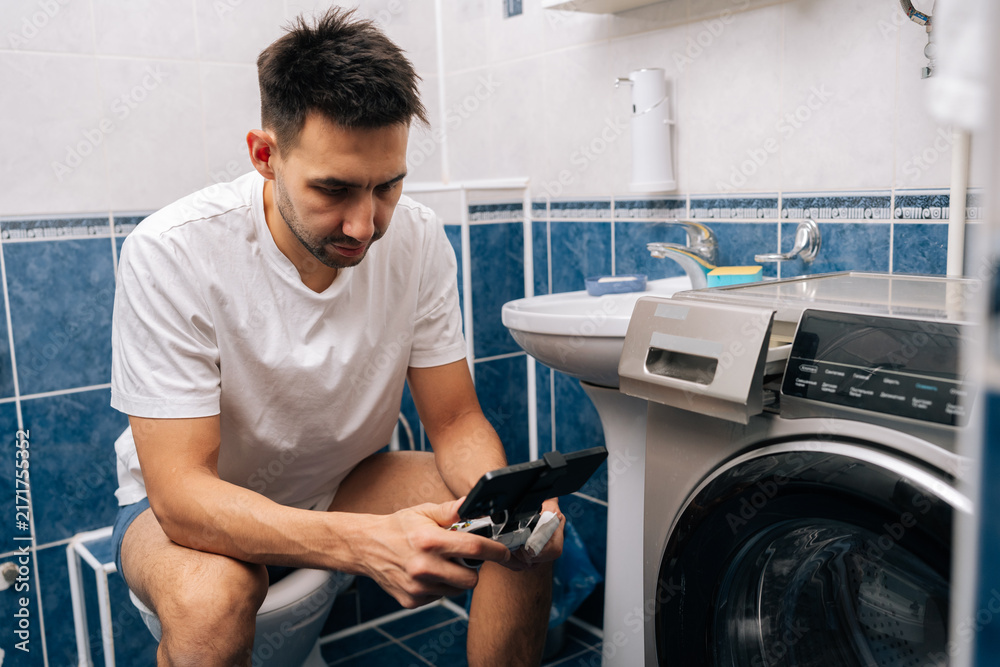Obraz premium Handsome bearded young adult man in white t-shirt focusing on mobile gaming using smartphone and gamepad while sitting on toilet, portraying modern habits and technology integration in daily life.