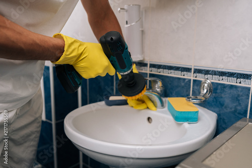 Konstfotografi Cropped shot of man wearing yellow gloves using power drill fitted with brush attachment to quickly scrub and sanitize bathroom sink and faucet for efficient home cleaning and maintenance, close-up