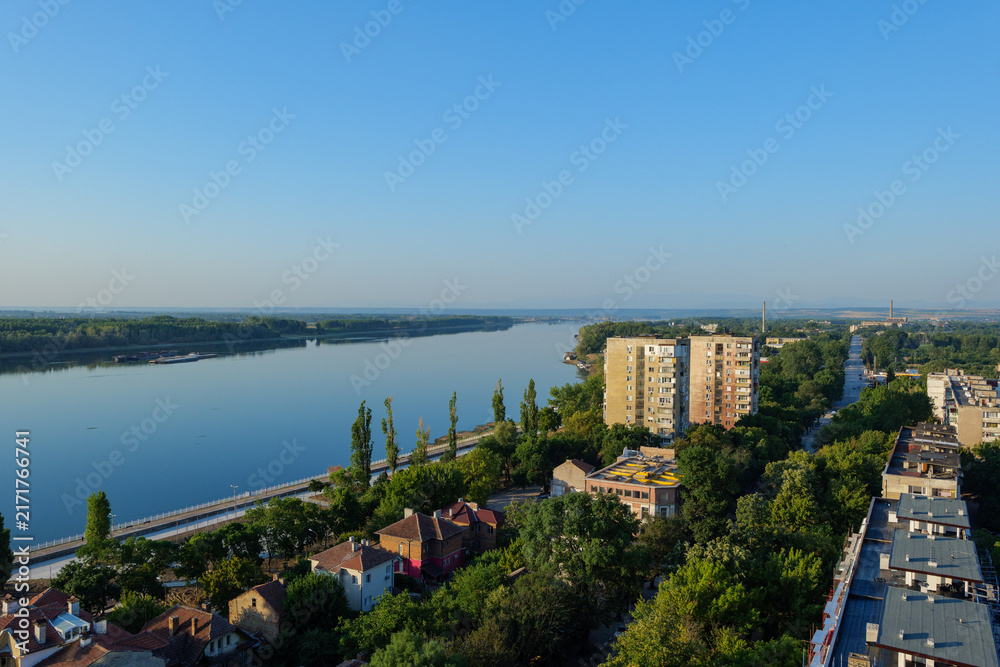 Naklejka premium Elevated panoramic view of Vidin, Bulgaria, featuring residential apartment blocks, a tree-lined boulevard and the urban landscape extending toward the horizon on a sunny summer day. Drone view