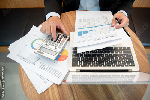 Businessman Reviewing Mortgage Calculator Documents with Calculator and Laptop on Wooden Desk
