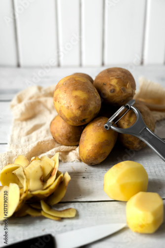 Konstfotografi Peeling raw potatoes with a knife and vegetable peeler on a white wooden table