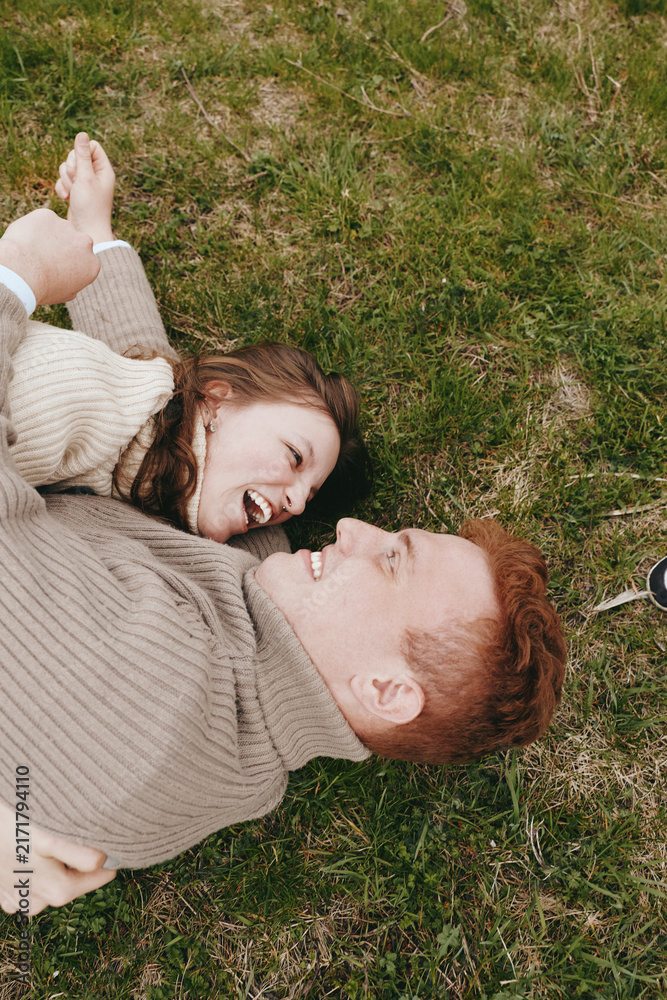 Naklejka premium Smiling couple lying on grass, outdoor daylight, and cozy sweaters convey warmth, affection, and relaxed leisure, creating a candid lifestyle moment filled with happiness and connection on a natural,