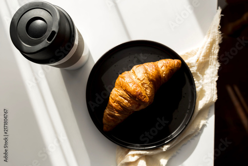  Fresh French croissant on a black plate and coffee in a reusable travel mug