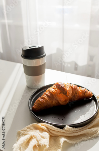  Fresh French croissant on a black plate and coffee in a reusable travel mug