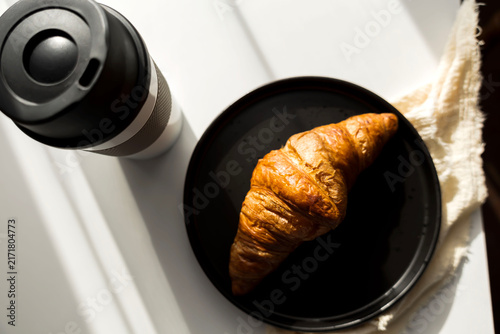  Fresh French croissant on a black plate and coffee in a reusable travel mug