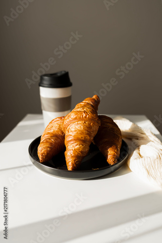  Fresh French croissant on a black plate and coffee in a reusable travel mug