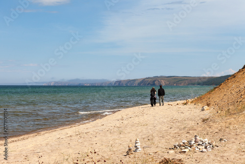 Baikal Lake in August. Sandy shore of Olkhon Island. Silhouettes of a couple of tourists strolling along the sandy beach of the Small Sea on a sunny day. Summer travel and outdoor recreation