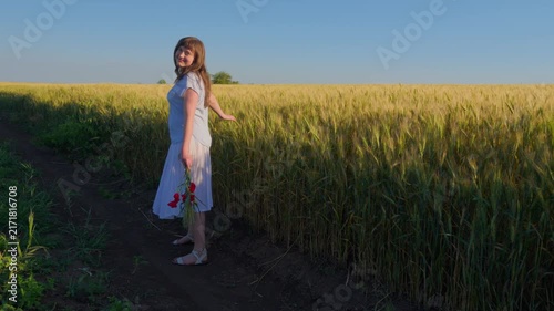 A young female teenager walking away along a path in a golden grain field while carrying a bouquet of red poppy flowers under a clear blue sky