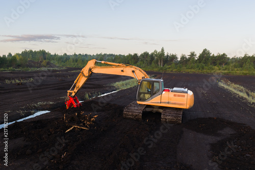 Fotografie Side view aerial drone shot of yellow heavy excavator machine working on peat bog field extracting soil near forest during sunrise in Estonia