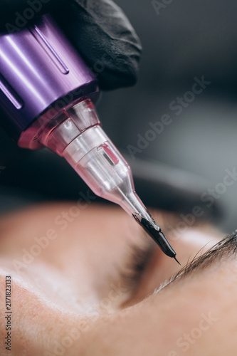 Macro close up of a permanent makeup needle with black pigment during an eyebrow tattoo procedure in a professional beauty studio.