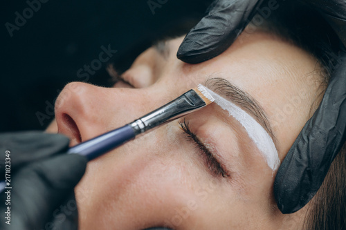 Permanent makeup artist applies white mapping paste to outline eyebrow shape before a cosmetic tattoo procedure in a professional beauty studio.