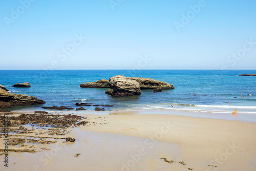 Foto Scenic View of Coastal Rock Formations, Sandy Shore, and Surfers in the Sea at B