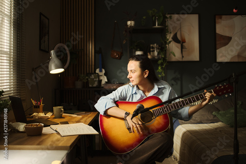 Bild auf Leinwand Young Caucasian woman playing acoustic guitar at home desk, looking at laptop screen