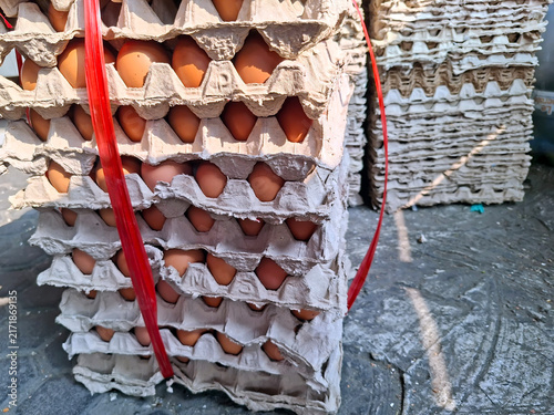 Foto A stack of brown chicken eggs stored in grey recycled paper pulp trays, tightly bound with red plastic strapping string outdoors