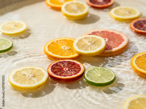 A highly realistic overhead lifestyle photograph of freshly sliced citrus fruits floating on the surface of clear, shallow water under bright natural sunlight
