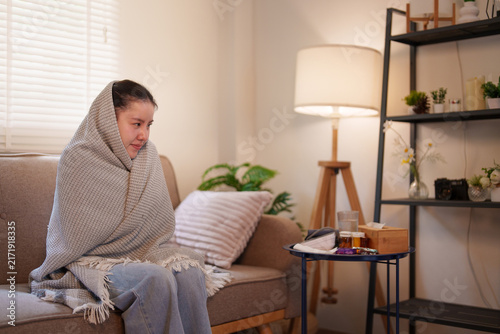 Sick Asian woman wrapped in blanket shivering with cold fever on couch at home