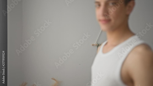 Young man touching his cheek with a hand and smiling in a bright studio by a window, wearing a white tanktop and casual pose; gentle serenity.