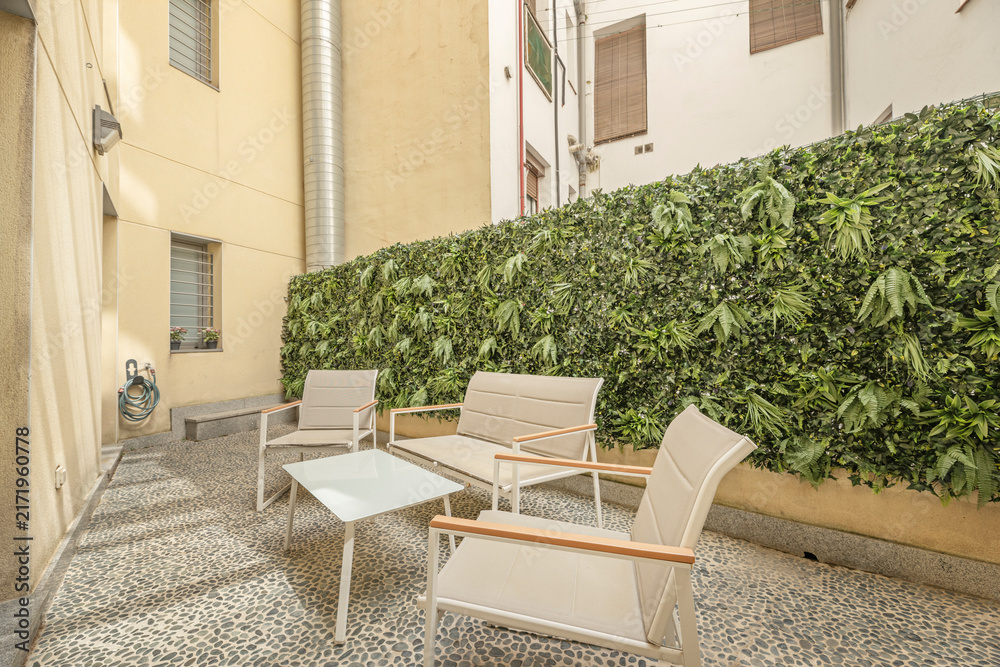 Fototapeta premium Sunlit inner courtyard with lounge chairs, sofa, and lush living plant wall between building facades.