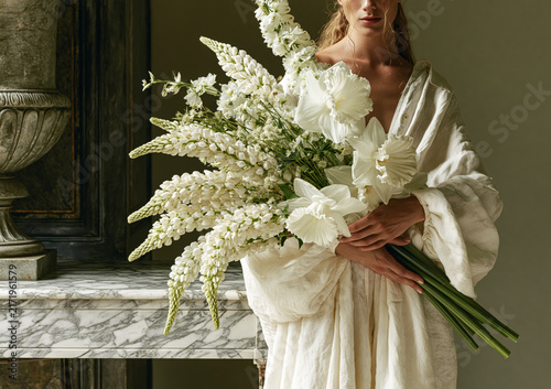 Woman in ivory dress holding a lush white floral bouquet in an ornate chateau interior.