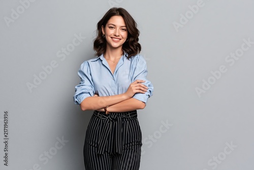 Smiling young woman with dark wavy hair wearing blue shirt and patterned trousers against gray