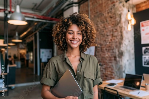 Smiling young woman with curly hair holding laptop standing in modern creative office interior