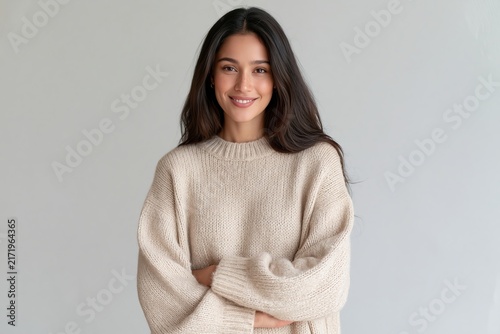 Young smiling woman with dark curly hair wearing beige knitted sweater posing against neutral wall